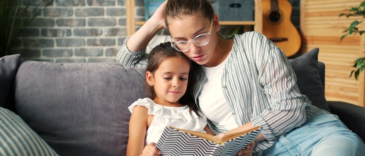 A parent and child reading a book together on a sofa in warm afternoon light